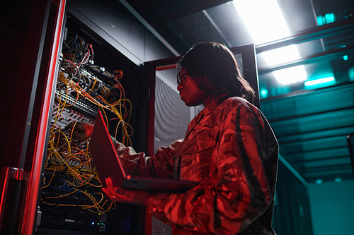 Low angle portrait of African-American woman managing computer network while working with supercomputer in server room lit by red light, copy space