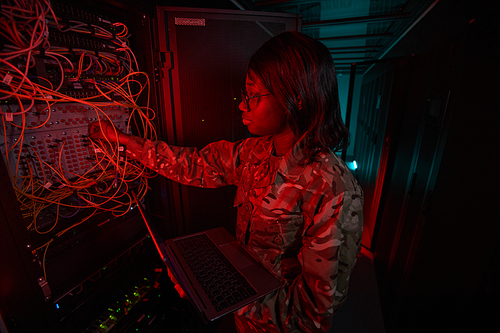 Side view at young African-American woman connecting cables while working with supercomputer in server room lit by red light, copy space