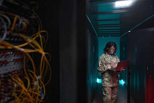 Portrait of young African-American woman wearing military uniform using laptop while standing in server room, copy space