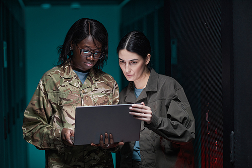 Waist up portrait of two young women wearing military uniform using laptop while standing in server room, copy space