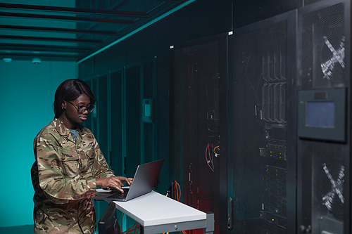 Waist up portrait of young African-American woman wearing military uniform using computer while setting up network in server room, copy space