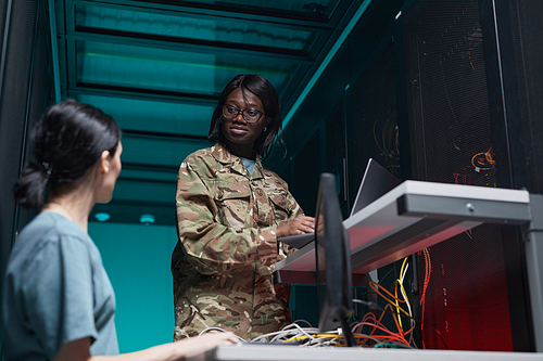 Low angle portrait of young African-American woman wearing military uniform using computer while setting up network in server room