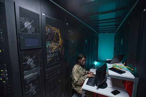 Wide angle portrait of young African-American woman wearing military uniform using computer while setting up network in server room, copy space