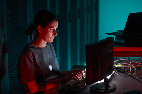 Side view portrait of female IT engineer using computer while working in server room lit by red light, copy space