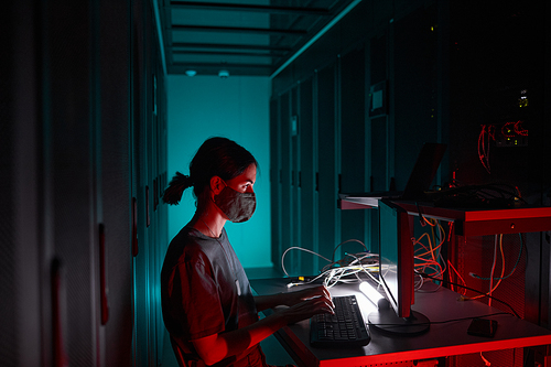 Side view portrait of female IT engineer wearing mask while using computer and working in server room lit by red light, copy space