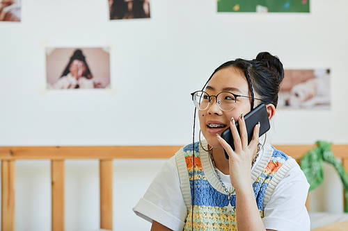 Portrait of Asian teenage girl speaking by smartphone while sitting on bed in cozy room, copy space