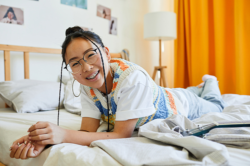 Portrait of carefree teenage girl laying on bed in cozy room and looking at camera