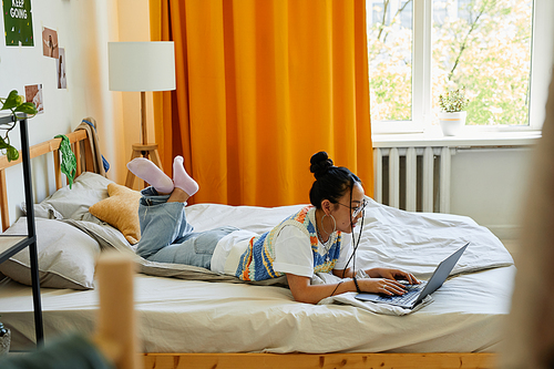 Full length portrait of carefree teenage girl using laptop on bed in cozy room, copy space