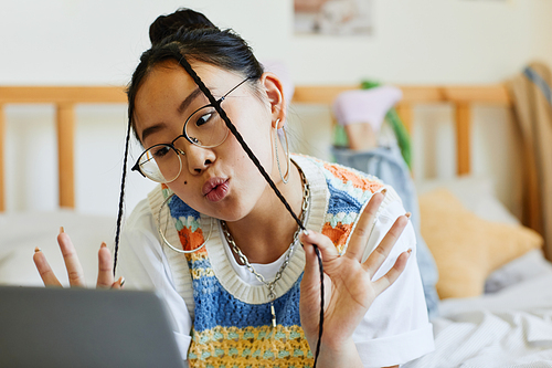 Portrait of teenage Asian girl using laptop on bed in cozy room interior and making cute faces to camera by video chat or livestream