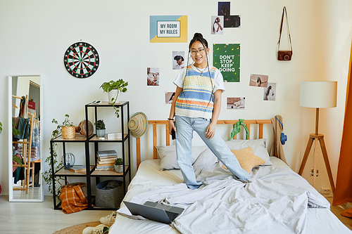 Full length portrait of teenage Asian girl standing on bed in cozy room interior and looking at camera, copy space