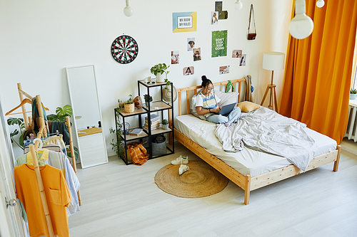 Wide angle portrait of teenage Asian girl using laptop while sitting on bed in cozy room interior, copy space