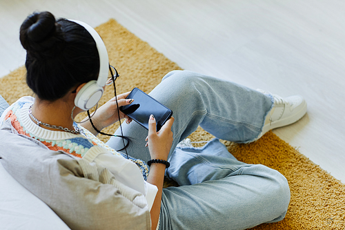 High angle view at trendy teenage girl using smartphone and listening to music while sitting on floor in cozy room, copy space