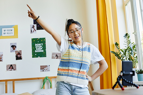 Waist up portrait of trendy teenage girl dancing to camera while filming videos in cozy room interior