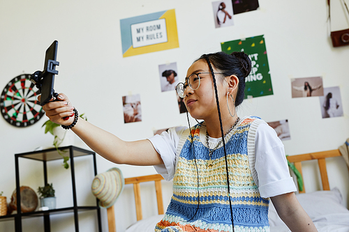 Side view portrait of trendy teenage girl holding smartphone while recording video or live streaming in cozy room