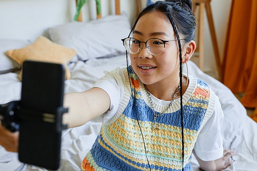 High angle portrait of trendy teenage girl holding smartphone while recording video or live streaming in cozy room