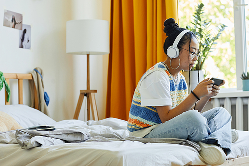 Side view portrait of trendy teenage girl playing mobile game vi smartphone while sitting on bed in cozy room, copy space