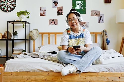 Full length portrait of trendy teenage girl looking at camera while sitting on bed in cozy room, copy space