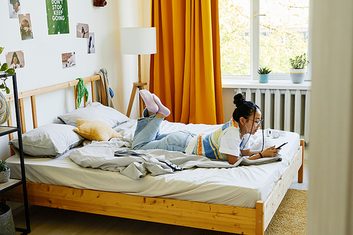 High angle portrait of trendy teenage girl laying on bed in cozy room and using smartphone, copy space