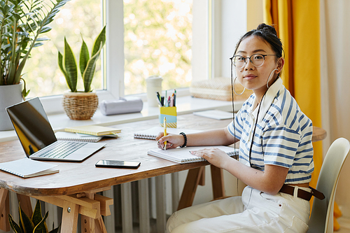 Portrait of Asian teenage girl studying at desk at home and looking at camera while writing in notebook, copy space