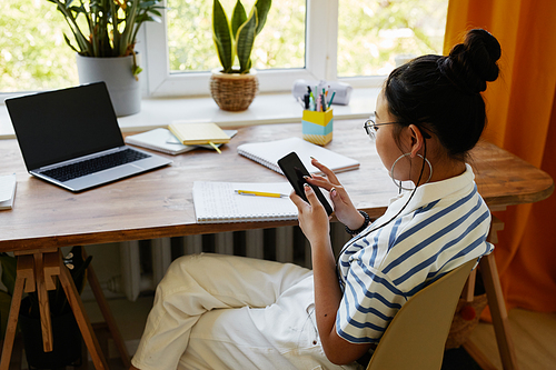 High angle portrait of teenage Asian girl using smartphone while sitting at desk at home, copy space