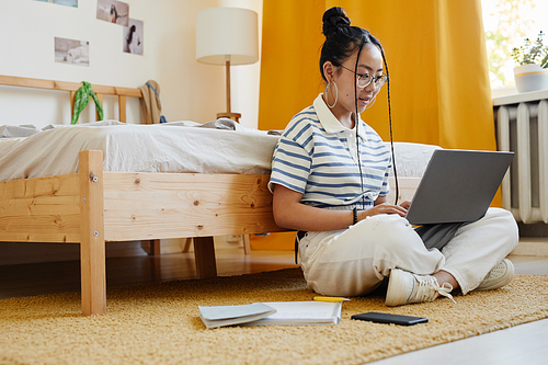 Full length portrait of Asian teenage girl using laptop while sitting on floor at home and studying, copy space