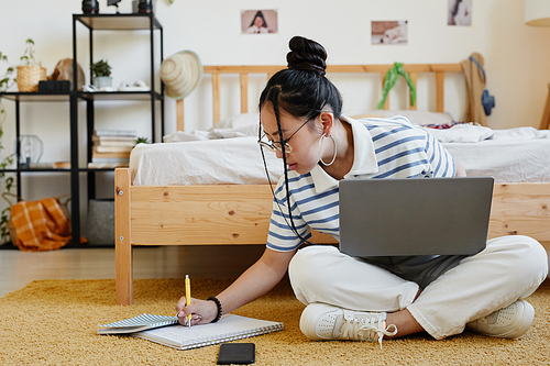 Full length portrait of Asian teenage girl using laptop while sitting on floor at home and doing homework, copy space