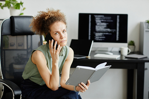 Serious businesswoman talking on mobile phone and planning her day while sitting on chair with notebook at office