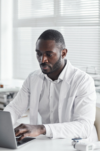 African young doctor in white coat sitting at the desk and working on laptop at his office