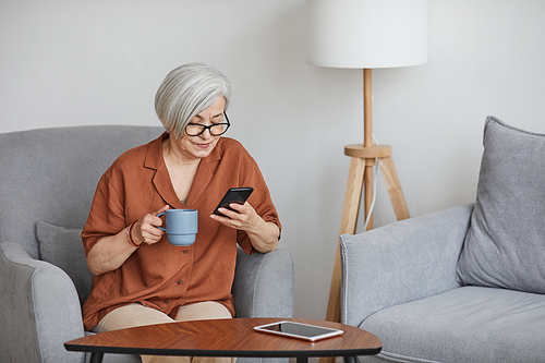 Minimal portrait of modern senior businesswoman using smartphone and enjoying coffee indoors, copy space