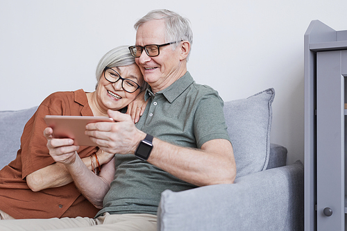 Minimal portrait of modern senior couple using tablet at home and smiling happily while sitting on couch, copy space