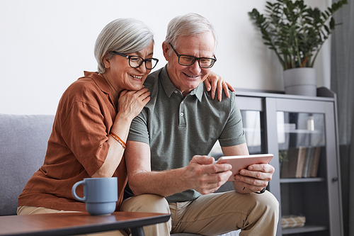 Minimal portrait of modern senior couple using tablet at home and smiling while sitting on couch together, copy space