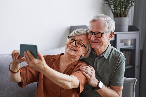 Portrait of happy senior couple taking selfie at home in minimal interior