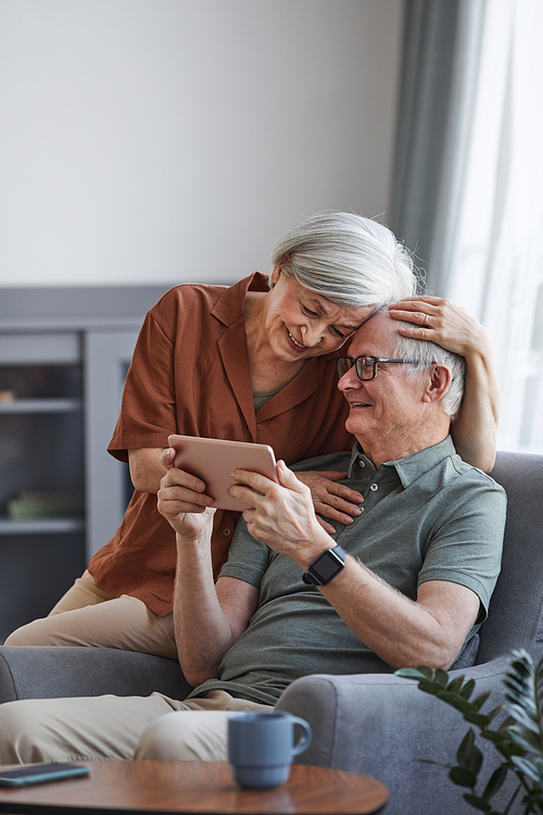 Vertical portrait of happy senior couple using digital tablet together at home and embracing