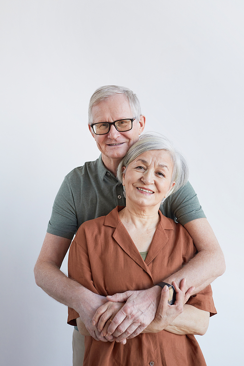 Vertical portrait of loving senior couple embracing and looking at camera while standing against white background