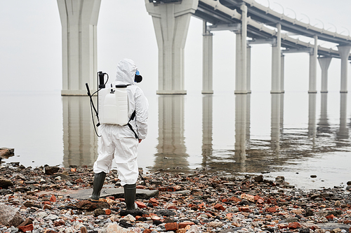 Full length back view at male worker wearing hazmat suit by water outdoors, industrial waste concept, copy space