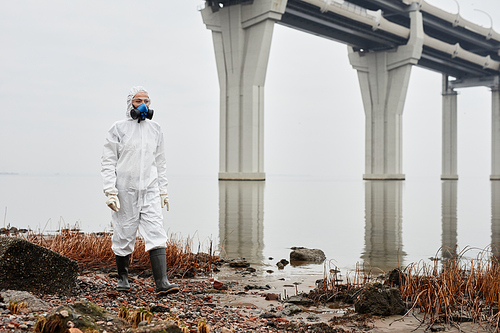 Full length portrait of male worker wearing hazmat suit walking by water outdoors, industrial waste concept, copy space