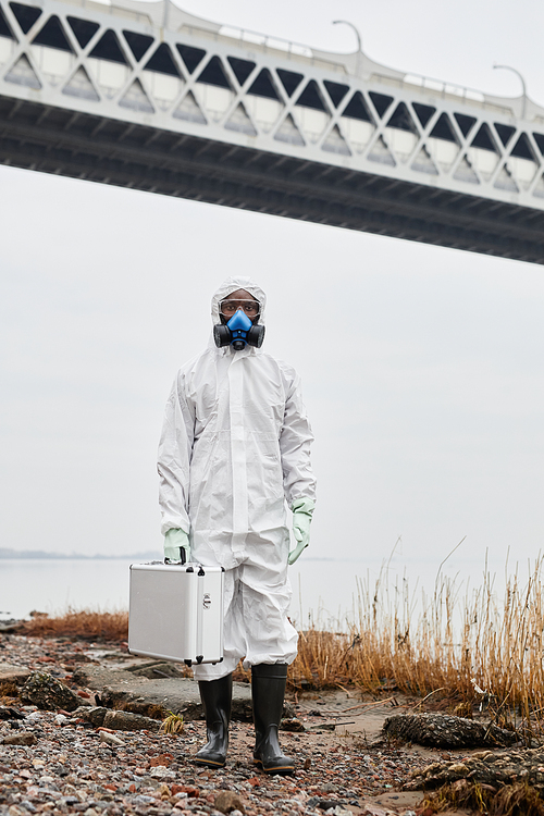 Vertical portrait of worker wearing hazmat suit walking by water outdoors and carrying samples case, industrial waste concept