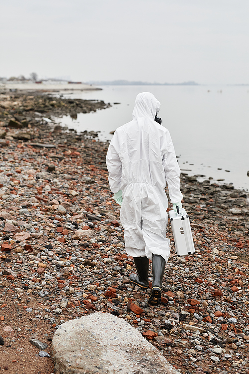 Vertical back view portrait of worker wearing hazmat suit walking by water outdoors and carrying samples case, industrial waste concept