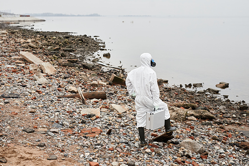 Full length back view portrait of worker wearing hazmat suit walking by water outdoors and carrying samples case, industrial waste concept, copy space