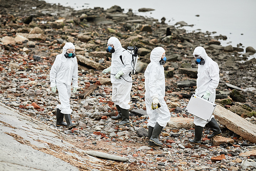 Group of workers wearing hazmat suits walking by water outdoors, toxic waste concept