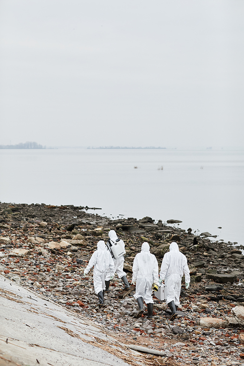 Vertical wide angle view at group of workers wearing hazmat suits walking by water outdoors , toxic waste concept, copy space