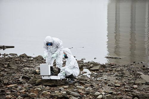 Two workers wearing hazmat suits collecting probe samples by water, toxic waste concept, copy space