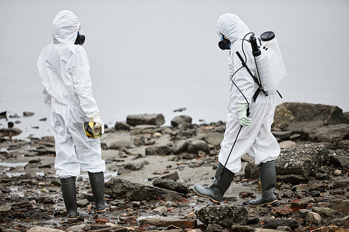 Full length portrait of two workers wearing hazmat suits collecting probe samples by water, toxic waste concept