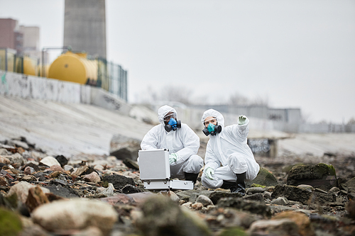 Full length portrait of two workers wearing hazmat suits collecting probe samples in industrial area, toxic waste concept