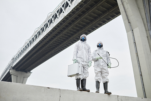 Industrial low angle portrait of two people wearing hazmat suits standing under bridge, copy space