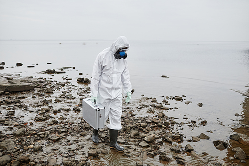 Full length portrait of man wearing hazmat suit collecting probes by water, toxic waste and pollution concept, copy space