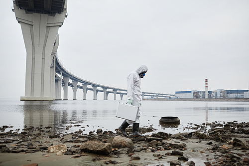Side view portrait of man wearing hazmat suit collecting probes by water, toxic waste and pollution concept, copy space