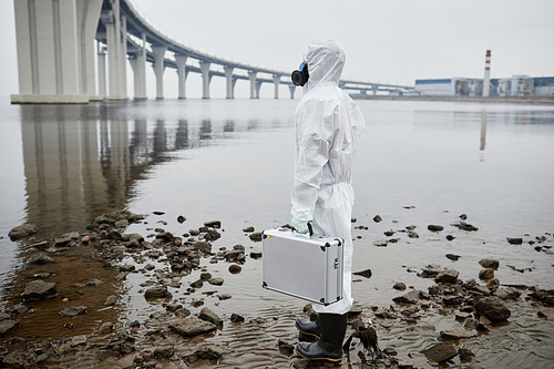 Full length portrait of man wearing hazmat suit standing by water, toxic waste and pollution concept, copy space