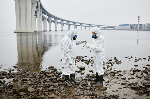 Full length portrait of two people wearing hazmat suit standing by water, toxic waste and pollution concept, copy space