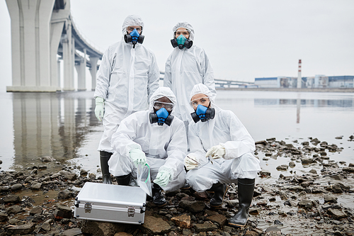 Group of people wearing hazmat suits looking at camera while standing by water, toxic waste and pollution concept, copy space
